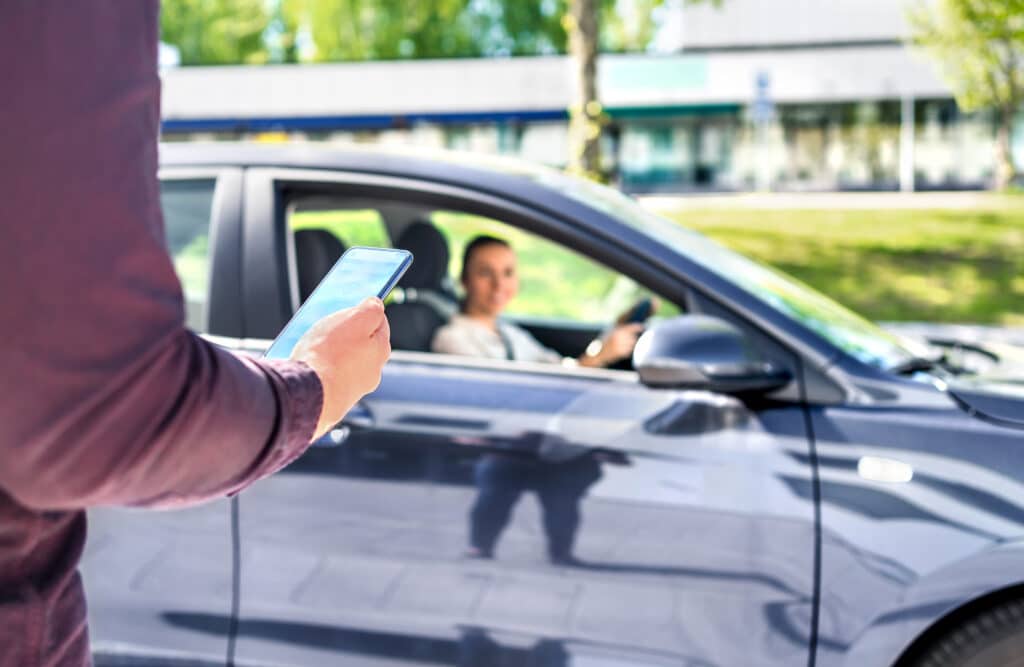 A person holding their phone in front of a car picking them up. Driving side hustle article.