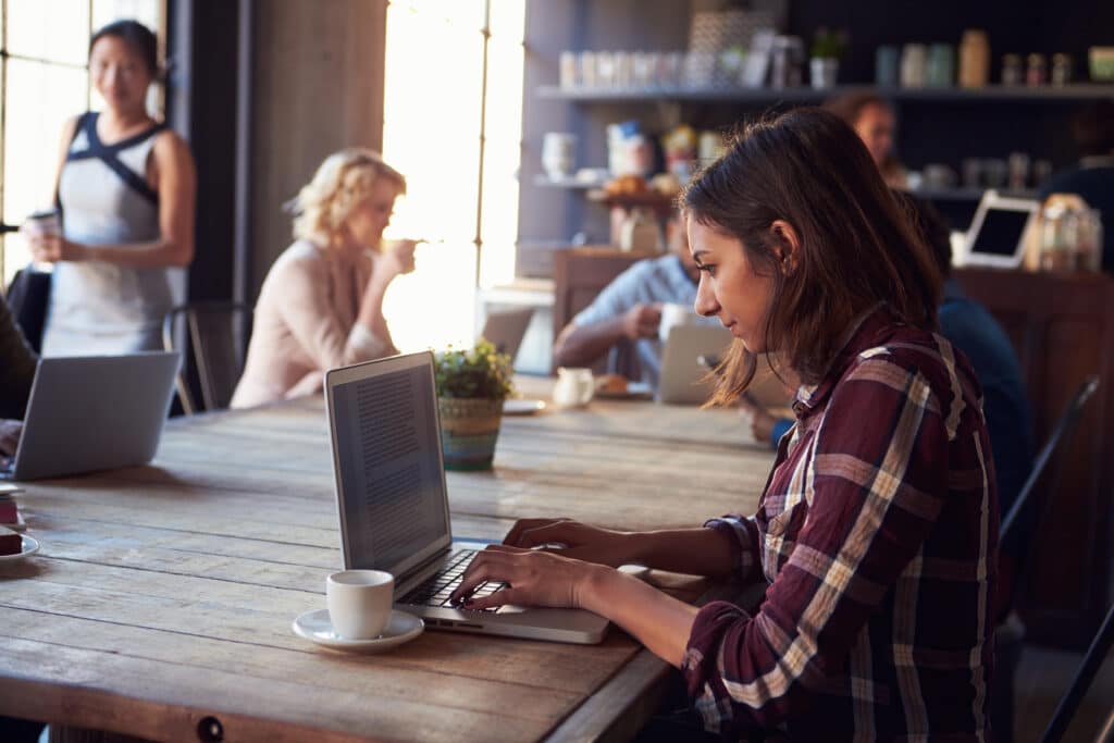 Woman with dark hair and a plaid shirt working on a laptop at a coffee shop.