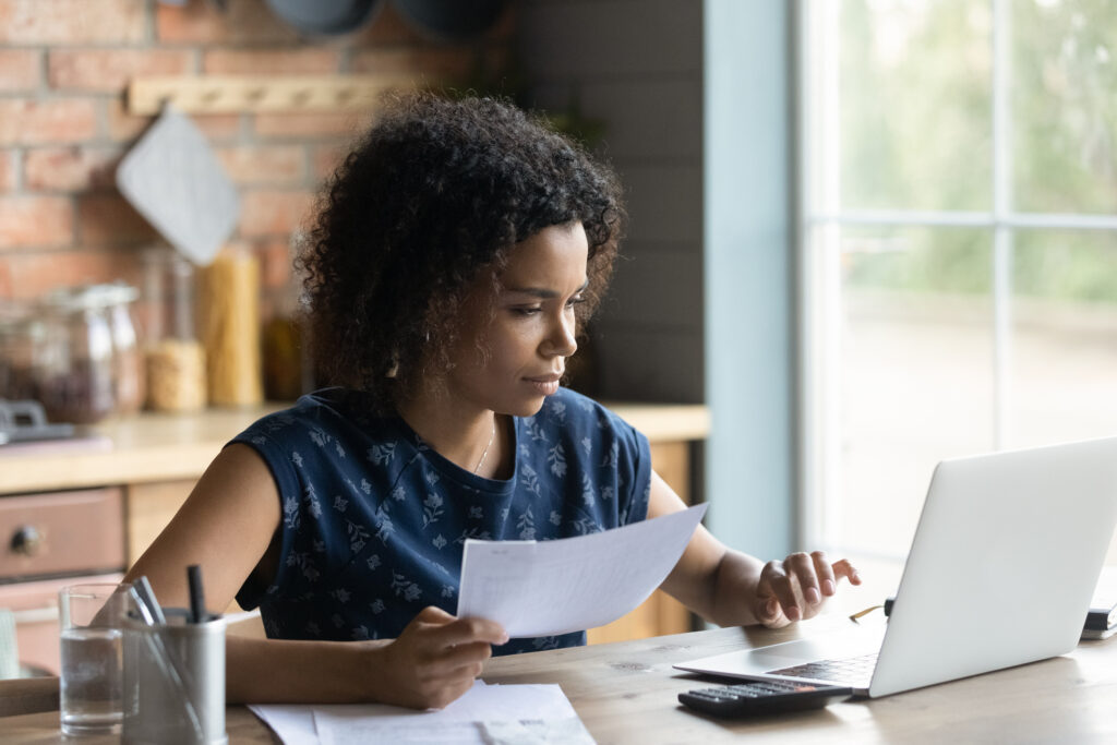 financial literacy. Serious millennial African American woman calculating taxes, budget, costs, checking paper invoice, receipt. Homeowner, tenant using laptop for paying bill, rent, mortgage, insurance fees