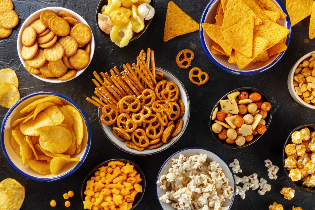 snack foods. Salty snacks, party mix. An assortment of crispy appetizers, overhead flat lay shot on a black background. Potato and tortilla chips, crackers, popcorn etc