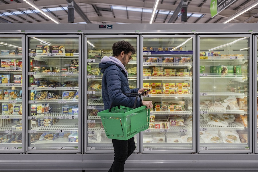 spending at the grocery store, man with green basket in frozen foods section looking at phone.